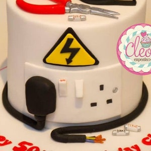 May include: A white cake decorated with an electrical theme. It features a yellow and black hazard sign, a plug, and a socket. Red scissors and a silver tool are on top. The cake is decorated with the words "Happy 50th Birthday Chris". A pink and white logo says "Cleo's cupcakes".