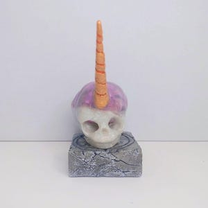 Puede incluir: Una escultura de calavera blanca con un cuerno de unicornio morado y dorado. La calavera se asienta sobre una base gris texturizada. El cuerno es de color naranja claro. La calavera tiene una sustancia morada y brillante.