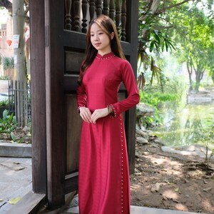 Könnte beinhalten: Eine Frau in einem langen, roten Kleid mit Perlenbesatz. Das Kleid hat einen hohen Kragen und lange Ärmel. Sie steht vor einer Holztür mit einem Gitterdesign.