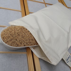 Könnte beinhalten: Ein cremefarbenes Kissen, teilweise gefüllt mit kleinen, braunen Körnern, mit einer weißen Schaufel, die mit denselben Körnern überquillt. Auf der Schaufel steht das Wort "spetex". Das Kissen liegt auf einem hellen, gitterförmigen Hintergrund.