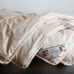 May include: A folded, cream-colored comforter with a quilted pattern. The comforter has a satin-like sheen and a tag with text. The comforter is folded over a gray surface. The tag reads "Papillon".