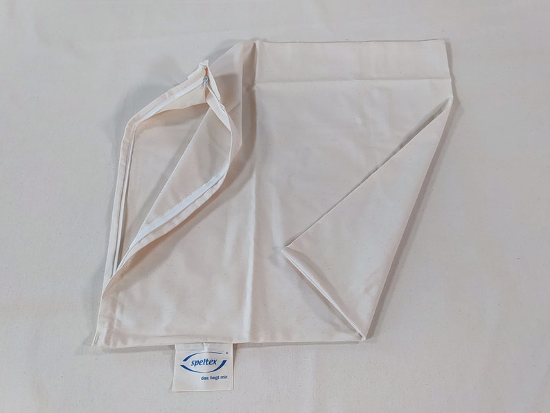 May include: A white pillowcase with a zipper closure. The pillowcase is folded in half and the zipper is open. The pillowcase has a blue label with the text "spertex das liegt mir" on it.