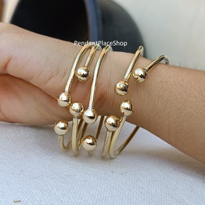 May include: Set of five gold-toned bangle bracelets with round ball accents. The bracelets are stacked on a person's wrist.
