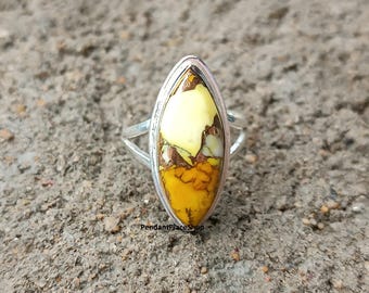 May include: A silver ring with an elongated, oval-shaped stone. The stone has yellow, brown, and white hues. The ring is set against a textured, grey surface. The text "PendantPlaceShop" is visible.