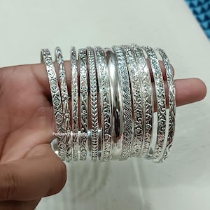 May include: A set of 12 silver bangles with intricate designs. The bangles are stacked on top of each other and have a variety of patterns, including floral, geometric, and wavy.