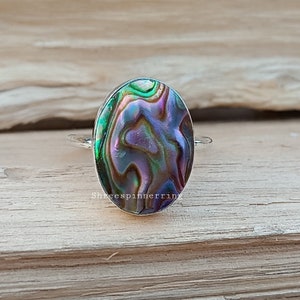Puede incluir: Un anillo de plata con una incrustación ovalada de concha de abulón. La concha tiene un patrón de remolino de verde, morado y marrón.