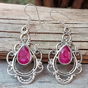 Peut inclure: Une paire de boucles d'oreilles argentées avec une pierre précieuse rose foncé en forme de goutte d'eau au centre. Les boucles d'oreilles présentent un design orné en filigrane avec une bordure torsadée et des accents décoratifs.