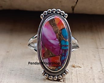 May include: Oval-shaped silver ring featuring a vibrant gemstone with a mix of colors: pink, orange, turquoise, and gold. The gemstone is set in a decorative bezel with a beaded edge. The ring has a split shank design.