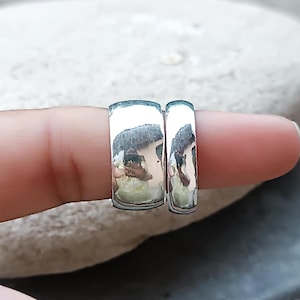 May include: Two polished silver rings, one slightly wider than the other, are displayed. The rings have a smooth, reflective surface, catching the light and reflecting the surroundings. The rings are likely for a couple.