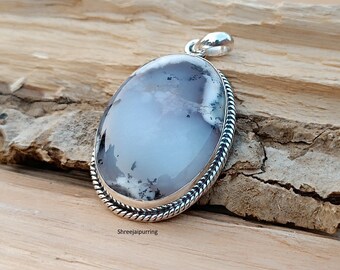 May include: Oval pendant with a light blue and white stone, set in a silver-colored metal frame with a rope-like border. The stone has a cloudy appearance with dark inclusions. The pendant is displayed on a wooden surface.