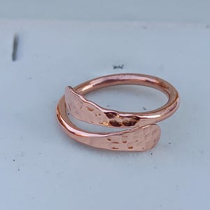 May include: A hammered copper ring with a unique wrap-around design. The ring is made of a single piece of copper that is bent and hammered to create a textured surface.