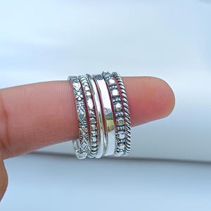 Puede incluir: Un conjunto de cinco anillos apilables de plata con diferentes texturas y patrones. Los anillos se llevan en un dedo.