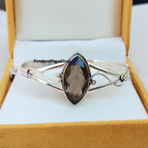 Könnte beinhalten: Ein silberfarbenes Armband mit einem großen, marquiseförmigen Rauchquarz-Edelstein. Das Armband hat zarte, wirbelnde Silberdesigns auf beiden Seiten des Steins. Das Armband wird in einer gelben Schachtel präsentiert. Der Text auf dem Armband lautet "PendantPlaceShop".