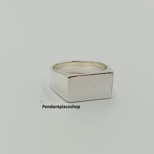 Könnte beinhalten: Ein polierter Siegelring aus Silber mit einer glatten, rechteckigen, flachen Oberseite. Der Ring hat eine massive Metallschiene und eine saubere, reflektierende Oberfläche. Der Text "Pendantplaceshop" ist in kleinen schwarzen Buchstaben abgedruckt.