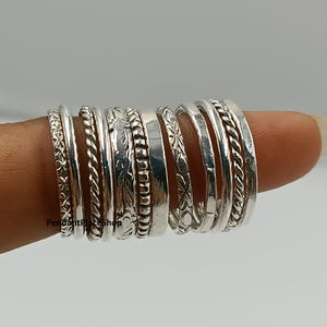 Może przedstawiać: Kolekcja srebrnych pierścionków o różnych wzorach, w tym plecionych, kwiatowych i teksturowanych. Pierścionki są ułożone jeden na drugim, prezentując różne style i szerokości. Pierścionki są prezentowane na palcu.