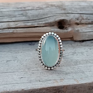 Pode incluir: Um anel de prata com uma pedra oval azul claro engastada em uma moldura de contas.