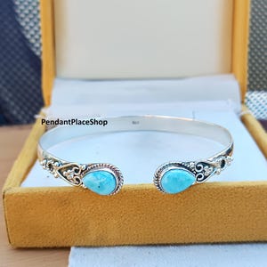 May include: A silver bangle bracelet with two turquoise stones set in the center of each side. The bracelet has a delicate floral design.