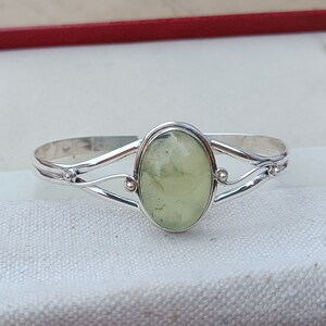 Puede incluir: Una pulsera de plata con una piedra ovalada verde engastada en el centro. La pulsera tiene un diseño delicado y ondulado.