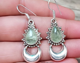 May include: A pair of silver earrings with green gemstone drops and crescent moon charms. The earrings have a delicate, bohemian style.