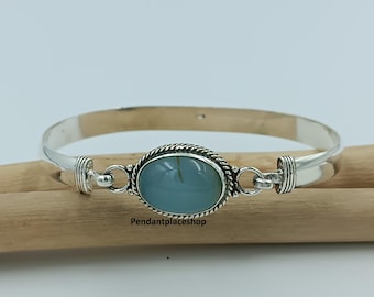 May include: A silver bangle bracelet with a light blue oval stone in the center. The stone is framed by a twisted rope design. The bracelet is resting on a wooden surface.