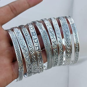 May include: A set of silver bangles with intricate engraved designs. The bangles are stacked on top of each other and feature various patterns, including floral motifs, geometric shapes, and braided designs.