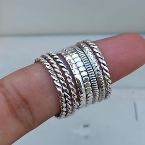 Puede incluir: Un conjunto de siete anillos apilables de plata con diferentes texturas, incluyendo trenzados, martillados y con cuentas.