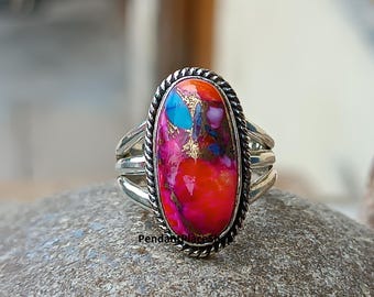 May include: Oval-shaped ring with a vibrant, multicolored stone. The stone features shades of red, pink, blue, and gold, set in a silver band with a braided border. The ring has a triple-band design.