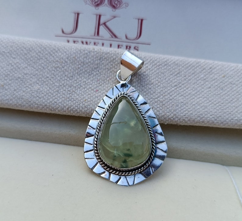 May include: A silver pendant featuring a green gemstone set within an intricate silver frame. The pendant is teardrop-shaped and has a silver bail for attaching a chain.