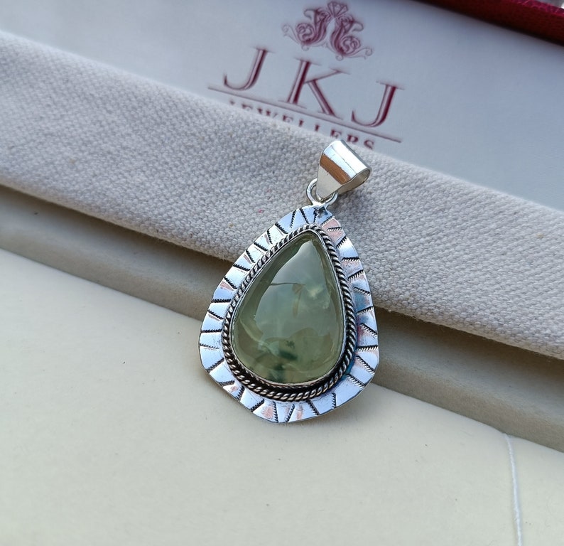 May include: A silver pendant featuring a green gemstone set in a teardrop shape. The pendant has an intricate silver border with a rope design.