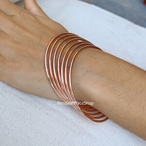 Puede incluir: Un conjunto de diez pulseras de oro rosa apiladas en la muñeca de una persona. Las pulseras son finas y tienen un acabado liso y pulido.