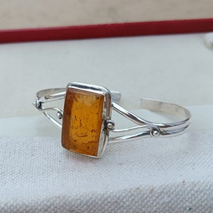 May include: A silver bangle bracelet featuring a rectangular amber stone set in the centre. The bracelet has a delicate design with two curved bands and small decorative beads.