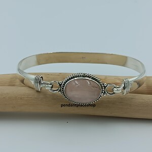 May include: A silver bracelet with an oval, light pink gemstone at the center. The gemstone is surrounded by a decorative rope-like border. The bracelet is resting on a wooden surface. The text "pendantplaceshop" is visible.