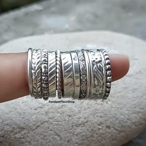 Puede incluir: Una colección de anillos de plata con diversas texturas y diseños, incluyendo bandas trenzadas, florales y estampadas. Los anillos están apilados en un dedo, mostrando los diferentes estilos y detalles. Los anillos son de plata.