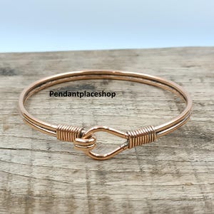 Puede incluir: Una pulsera brazalete de color cobre con cierre de gancho. La pulsera presenta un diseño sencillo con detalles de alambre enrollado alrededor del gancho y a ambos lados del cierre. El texto "Pendantplaceshop" es visible.