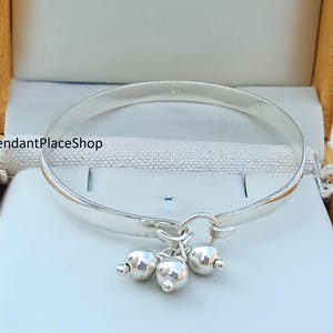 Puede incluir: Un brazalete de plata con un dije de tres bolas de plata colgando de él. El brazalete está en una caja de terciopelo amarillo.