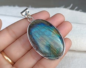 Puede incluir: Pendiente de plata con una gran piedra de labradorita de forma ovalada con reflejos iridiscentes azules, verdes y dorados. El pendiente tiene una anilla de plata.