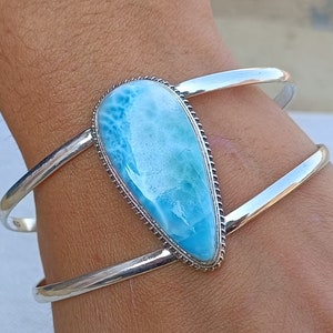 Könnte beinhalten: Ein silbernes Armreif mit einem großen, tropfenförmigen blauen und weißen Edelstein. Der Edelstein hat ein wirbelndes, marmoriertes Muster.