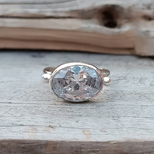 Op de afbeelding: Een zilveren ring met een grote, heldere ovale edelsteen. De ring heeft twee kleine zilveren balletjes aan weerszijden van de edelsteen.