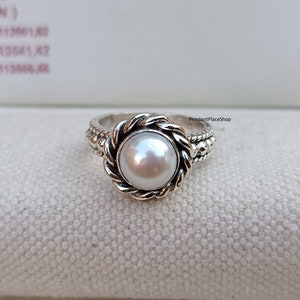Op de afbeelding: Een zilveren ring met een witte parel gezet in een touwachtig ontwerp. De ring ligt op een witte stoffen achtergrond. PendantPlaceShop staat in de rechterbovenhoek van de afbeelding.