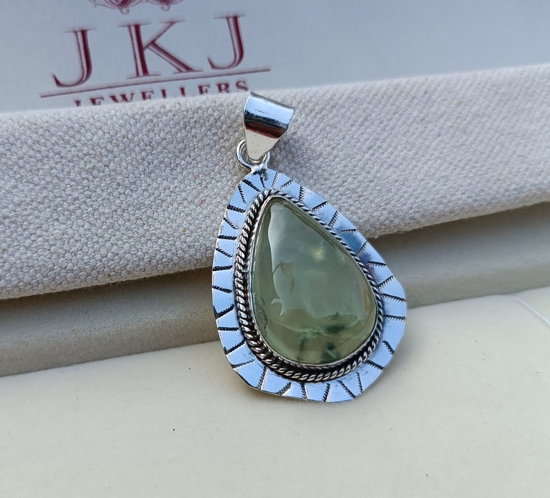 May include: A silver pendant featuring a green gemstone set in a teardrop shape. The pendant has a detailed silver border with a rope-like design.