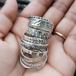 Peut inclure: Une pile de bagues en argent avec divers motifs, dont des motifs floraux, tressés et texturés. Les bagues sont tenues dans une main, mettant en valeur les différents styles et textures. Les bagues sont probablement pour les doigts et sont en métal brillant.
