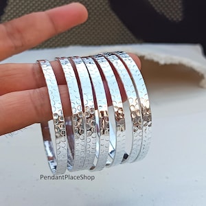 Peut inclure: Ensemble de sept bracelets argentés avec une texture martelée. Les bracelets sont empilés, reflétant la lumière. Le nom de la boutique "PendantPlaceShop" est visible en bas de l'image.