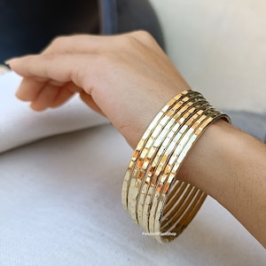 Può includere: Un set di otto braccialetti dorati con una texture martellata. I braccialetti sono impilati sul polso di una persona.