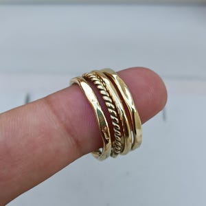 May include: Four gold rings stacked on a finger. The rings are of different widths and styles, including a hammered ring and a rope-style ring.