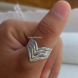 以下が含まれることがあります： V字型のデザインのシルバーリング。リングは3つのバンドが重ねられて作られています。リングは親指に着用されています。