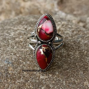 May include: A silver ring featuring two teardrop-shaped stones. The stones are a deep red with gold flecks and white and pink accents. The ring has a braided border and a split shank design.