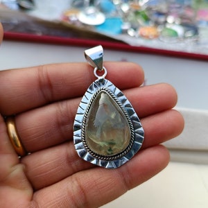 May include: A silver pendant featuring a green and white gemstone in a teardrop shape. The pendant has an intricate silver border.