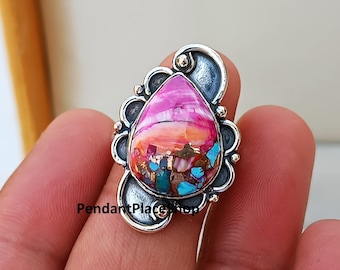 May include: A silver ring with a pink, orange, and turquoise gemstone set in a teardrop shape. The ring has a decorative, ornate design.