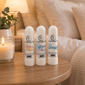 May include: Three white aromatherapy sticks with the words "Rise & Shine", "Stress Ease", and "Breathe" printed on them. The sticks are on a wooden table next to a lit candle and a small plant. The background includes a bed and a lamp.