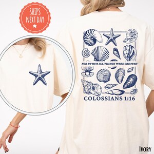 May include: Ivory-colored t-shirt featuring a navy blue starfish on the front and a collection of navy blue seashells with the text "FOR BY HIM ALL THINGS WERE CREATED COLOSSIANS 1:16" on the back. The shirt has a round neck.